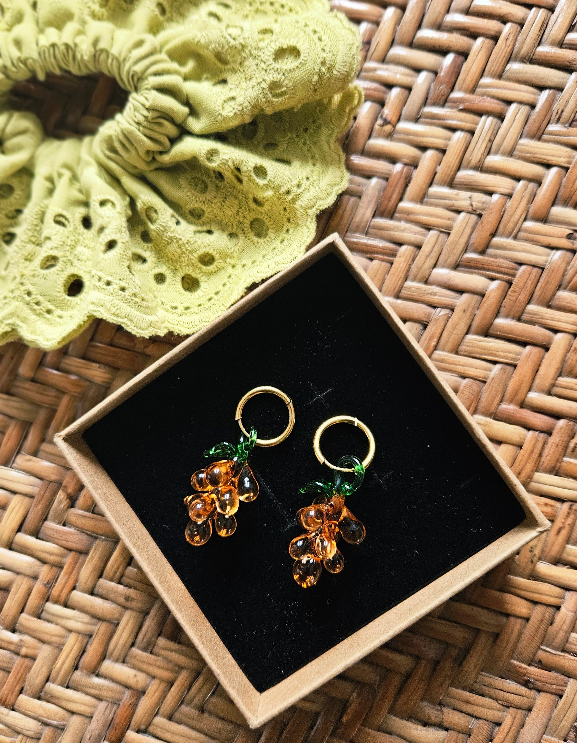 Pair of earrings in a box on a woven surface with a green scrunchie.