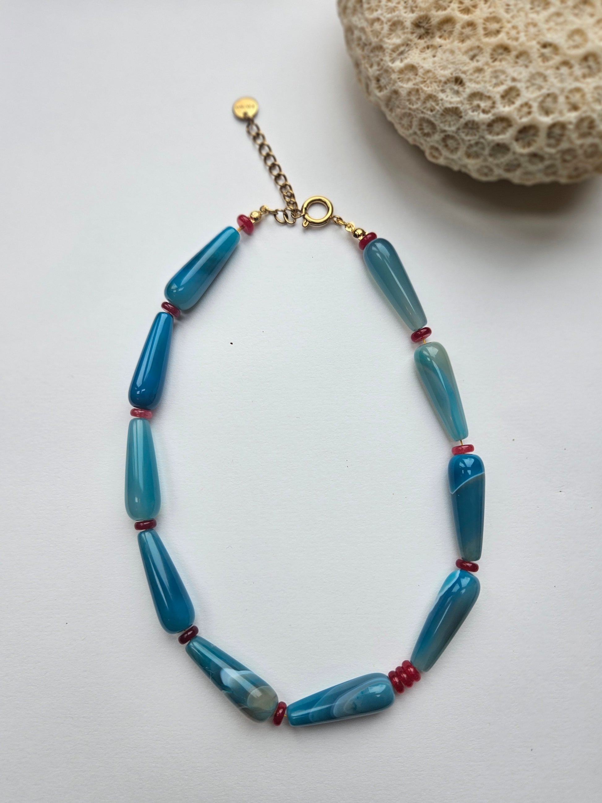 Blue beaded necklace with red accents on a white surface next to a piece of coral.